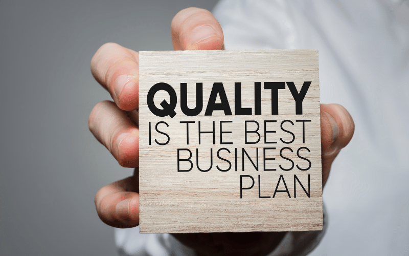 A hand holding a wooden block printed with the words "Quality is the best business plan"