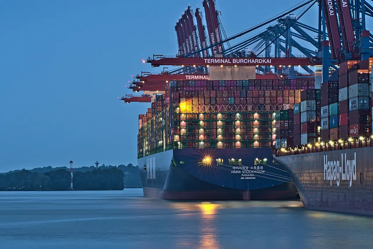 A loaded container ship berthed under gantry cranes at dusk